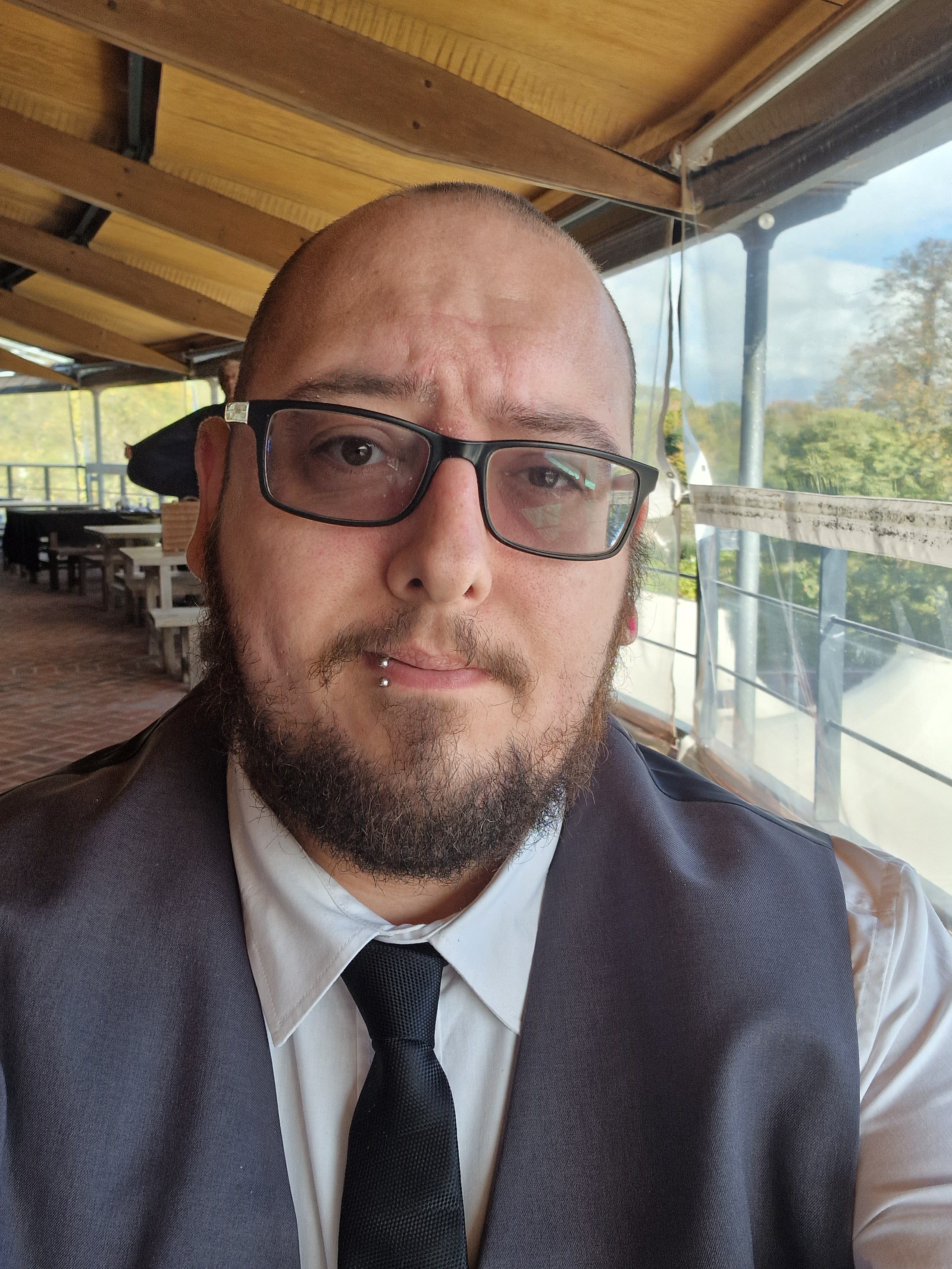 Image of David. A white, Mediterranean, masculine person with a shaved head, thick rimmed glasses, lip piercing, and an ear tunnel. David is wearing a waistcoat, white shirt, and tie. He is sat at a covered picnic table.